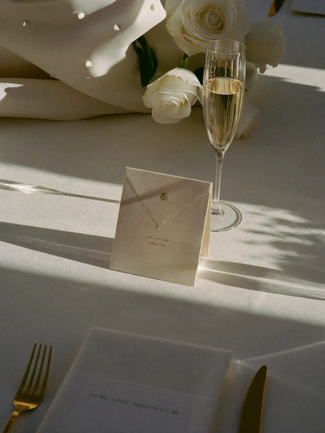 Elegante cremefarbene Hochzeitspapeterie zwischen weißen Rosen und goldenem Besteck